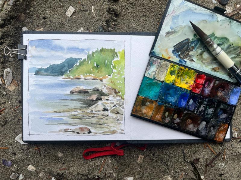 A sketchbook on the sand with a watercolor palette and paint brush overlaying it.