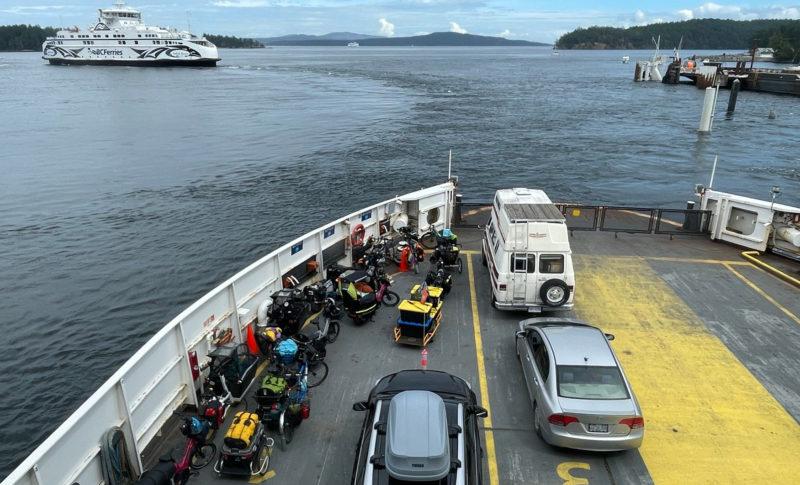 Many cargo bicycles parked on the left side of a ferry car deck with a view of the water and another ferry in the distance.