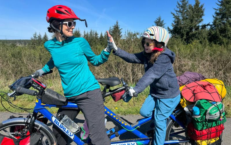 Two people wearing helmets and straddling a tandem bicycle giving each other a high give.