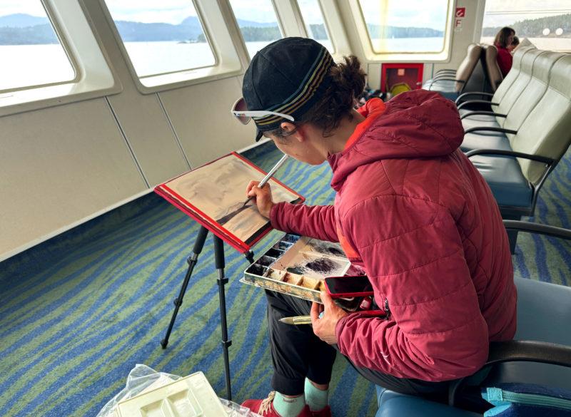A woman in a hat and jacket holds a paint brush over a tripod with a paper and board on it while on a ferry.